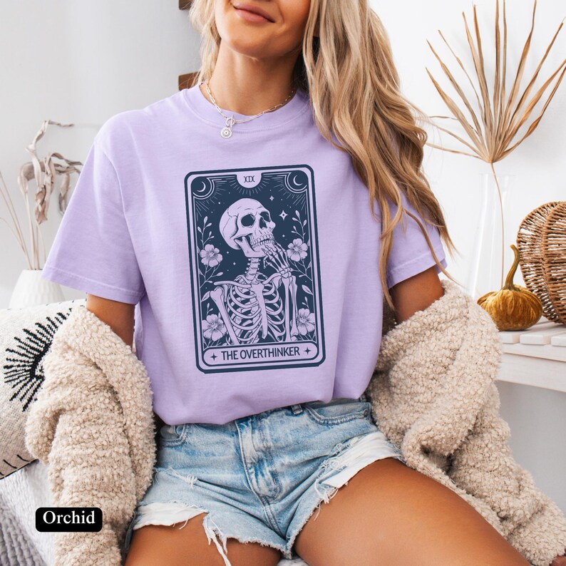 May include: A lavender t-shirt with a tarot card design featuring a skeleton with the text "The Overthinker".