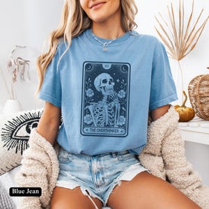 May include: A light blue t-shirt with a graphic of a skeleton holding a flower and the text "The Overthinker".