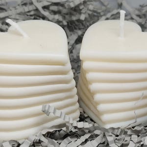 Puede incluir: Dos velas blancas en forma de corazón apiladas una encima de la otra. Las velas están hechas de múltiples capas de cera, creando un efecto de capas.