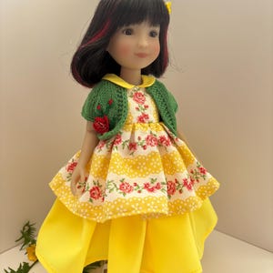 May include: A doll in a yellow dress with a floral bodice and a green knitted bolero. The doll has black hair with red highlights and a yellow flower. The dress features a layered skirt and white shoes.