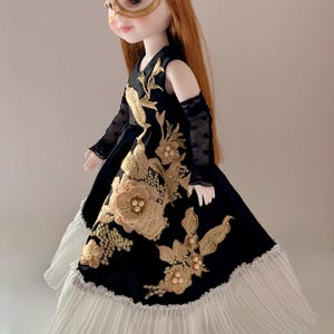May include: A doll with long reddish-brown hair wearing a black dress with gold floral embroidery, pearl accents, and a white pleated hem. The doll wears a gold lace mask with sequins and black mesh arm sleeves, showcasing intricate detailing.