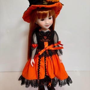 May include: A doll in a Halloween witch costume. The doll has long red hair and wears a black witch hat with orange trim and flowers. The orange dress has black lace trim and a black bodice. The outfit is completed with black boots.