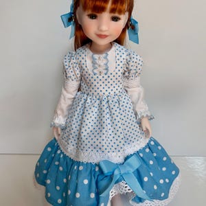 Peut inclure: Poupée aux cheveux roux coiffés en couettes, ornées de nœuds bleus. Elle porte une robe blanche et bleue à pois, bordée de dentelle et ornée d'un nœud bleu à la taille. La robe a des manches longues et un col à volants.