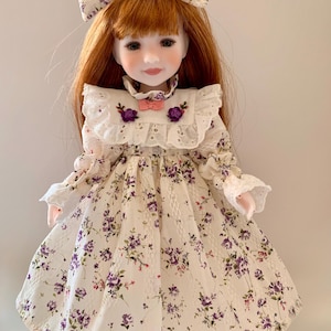 May include: A doll with red hair and a white dress decorated with purple floral patterns. The dress has a ruffled collar, lace details, and a matching bow in the hair. The doll's face is delicately painted with a soft expression.