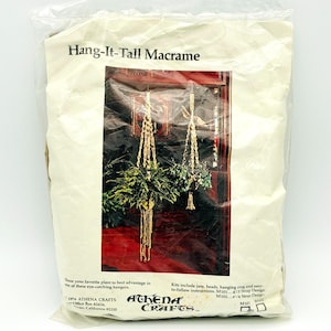 May include: A plastic-wrapped craft kit titled Hang-It-Tall Macrame by Athena Crafts. The packaging features an image of two beige macrame plant hangers holding green plants, with text detailing kit contents like jute and beads.
