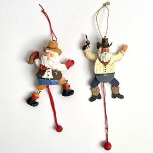 May include: Two hanging ornaments featuring figures dressed in cowboy attire. The left figure wears a tan hat, brown vest, and blue pants with red accents. The right figure wears a dark hat, cream shirt, and blue pants, holding a small black pistol.