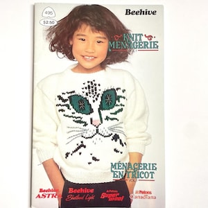 May include: A young person wearing a white sweater with a cat face design. The sweater is part of a knitting pattern book titled "Knit Menagerie" by Beehive. The book cover also features the text "Ménagerie en Tricot".