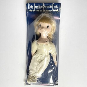 May include: A Cache Junction porcelain doll in its original packaging. The doll has a painted face with blonde hair and is wearing a cream-colored outfit. The packaging is blue with white text.