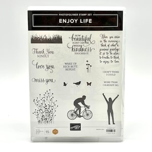 May include: A photopolymer stamp set titled ENJOY LIFE featuring various black ink designs including a cyclist, a person with arms raised, grass, flowers, butterflies, a bird, and several inspirational phrases in different fonts on a white sheet.