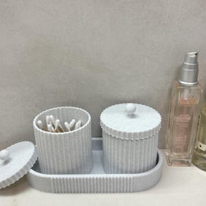 Op de afbeelding: Twee witte keramische containers met deksels, een gevuld met wattenstaafjes, op een witte dienblad. Twee flessen vloeibare zeep, een heldere en een amberkleurige, staan op het aanrecht achter de containers.