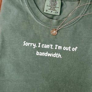 Puede incluir: Una camiseta verde musgo Comfort Colors con el texto "Sorry, I can't. I'm out of bandwidth." Un collar dorado con un colgante descansa sobre la camiseta. La camiseta está doblada sobre una superficie blanca, con un sombrero marrón y una planta seca en el fondo.