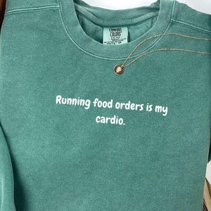 May include: A light green sweatshirt with the text "Running food orders is my cardio." in white. The sweatshirt has a crew neck and long sleeves. A gold necklace with a pendant is visible. The words "Sweatshirt" and "Light Green" are also in the image.