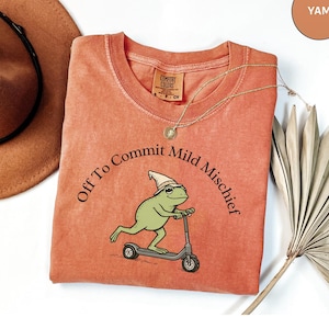May include: A yam-coloured Comfort Colors t-shirt featuring the text "Off To Commit Mild Mischief" around a cartoon frog on a scooter. The frog wears a pointed hat. A gold necklace and a brown hat are visible.