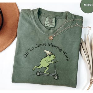 May include: Moss green t-shirt with the text "Off To Chase Missing Work" and a cartoon frog riding a scooter. The shirt has a crew neck and short sleeves. A gold necklace and a brown hat are also in the image.