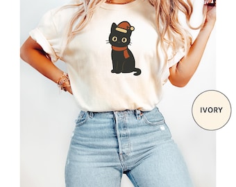 Puede incluir: Camiseta de color marfil con un gato negro de dibujos animados que lleva un gorro de Papá Noel y una bufanda roja. El gato tiene ojos grandes y redondos y está sentado erguido. La camiseta se combina con vaqueros azules. La palabra "IVORY" está en un círculo a la derecha.