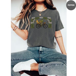 Puede incluir: Camiseta gris oscuro con una ilustraci&oacute;n caprichosa de dos ranas montando en bicicleta bajo un arco floral. El dise&ntilde;o es en tonos verdes, amarillos y blancos. La camiseta tiene un corte holgado y cuello redondo.