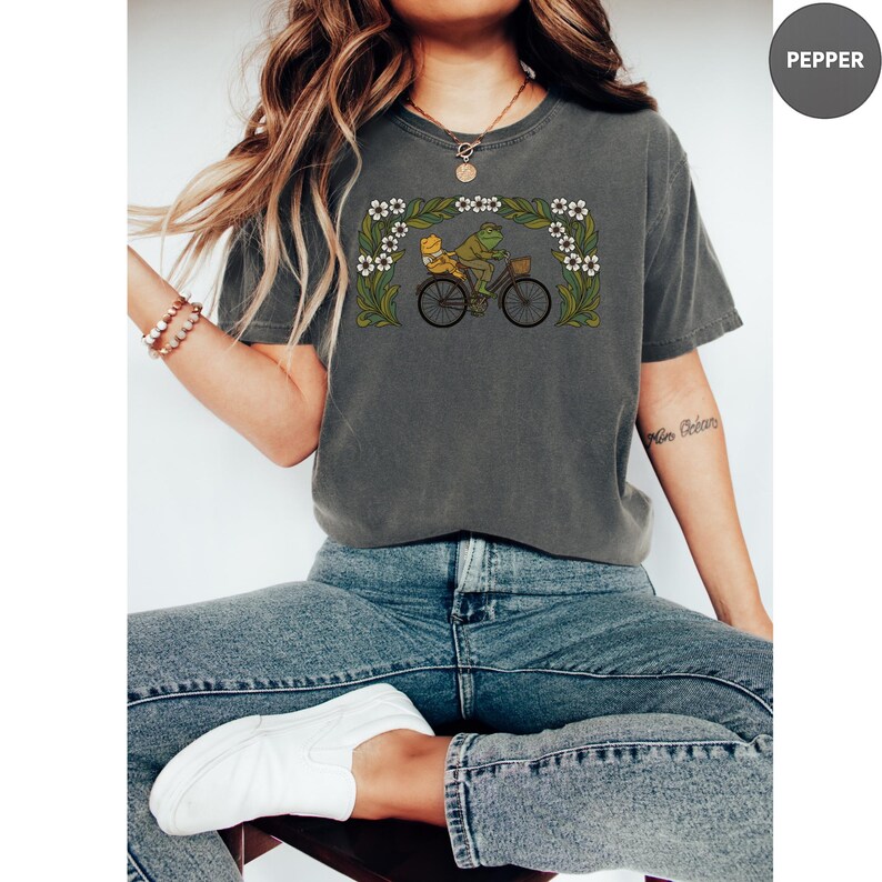 Puede incluir: Camiseta gris oscuro con una ilustraci&oacute;n caprichosa de dos ranas montando en bicicleta bajo un arco floral. El dise&ntilde;o es en tonos verdes, amarillos y blancos. La camiseta tiene un corte holgado y cuello redondo.