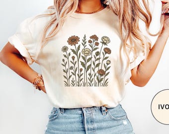 May include: A cream-colored short-sleeve t-shirt featuring a central graphic print of various wildflowers in shades of brown, tan, and muted green. The design depicts a collection of stems, leaves, and blossoms arranged in a vertical cluster.