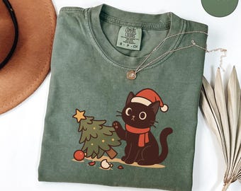 Op de afbeelding: Een mosgroen Comfort Colors T-shirt met een zwarte kat met een kerstmuts en rode sjaal, die een kerstboom versiert. Een gouden ketting en een bruine hoed zijn ook zichtbaar. Het shirt heeft de tekst "COMFORT COLORS".