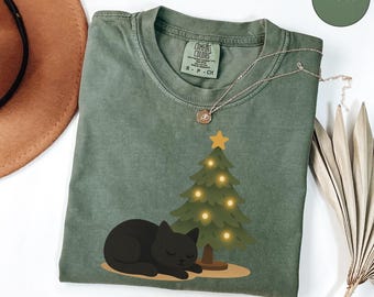 Puede incluir: Camiseta verde musgo con un gato negro durmiendo junto a un árbol de Navidad con luces amarillas. La camiseta tiene cuello redondo y mangas cortas. Un collar dorado está drapeado sobre la camiseta. La etiqueta dice "Comfort Colors".