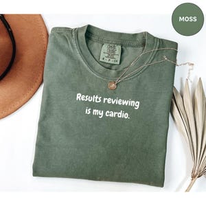 Pode incluir: Uma camiseta verde musgo com a frase branca "Results reviewing is my cardio". A camiseta tem gola redonda e mangas curtas. Um colar de ouro com um pingente repousa sobre a camiseta. A camiseta está dobrada sobre uma superfície branca.
