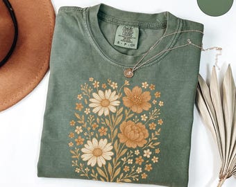 May include: A moss green short-sleeve t-shirt featuring a central graphic print of various flowers in shades of cream, tan, and brown. The shirt has a crew neckline and a Comfort Colors brand label on the inside collar with size S-P-CH visible.