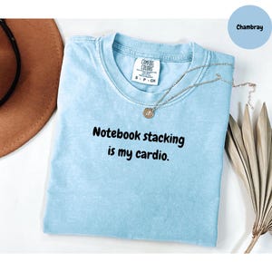 以下が含まれることがあります： 「Notebook stacking is my cardio」のテキストが入った水色のTシャツ。ペンダント付きのシルバーネックレスがシャツの上にあります。「Comfort Colors」のブランドで、サイズはSです。