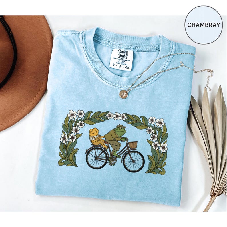 Puede incluir: Camiseta Comfort Colors azul claro con una ilustraci&oacute;n caprichosa de dos ranas montando en bicicleta a trav&eacute;s de un arco floral. El dise&ntilde;o incluye una rana con sombrero y chaqueta, y la camiseta tiene una etiqueta de talla.