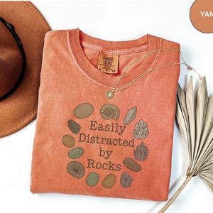 以下が含まれることがあります： クルーネックのヤム色のTシャツで、ダークブラウンのフォントで「Easily Distracted by Rocks」というテキストの周りに様々な岩や結晶が円状に配置されたグラフィックプリントが施されています。Comfort Colorsのラベルが見えます。
