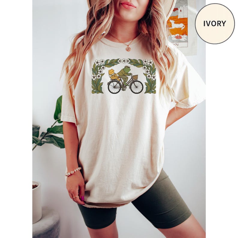 Puede incluir: Camiseta color marfil con un gr&aacute;fico caprichoso de una rana y un pato montando en bicicleta, enmarcado por un borde floral. El dise&ntilde;o es en tonos verdes, amarillos y negros. La camiseta es de corte holgado.
