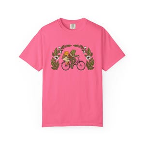 Puede incluir: Camiseta rosa brillante con una ilustraci&oacute;n caprichosa de dos ranas montando en bicicleta. Las ranas est&aacute;n rodeadas por un arco floral con flores blancas y hojas verdes. La camiseta es de color rosa s&oacute;lido.