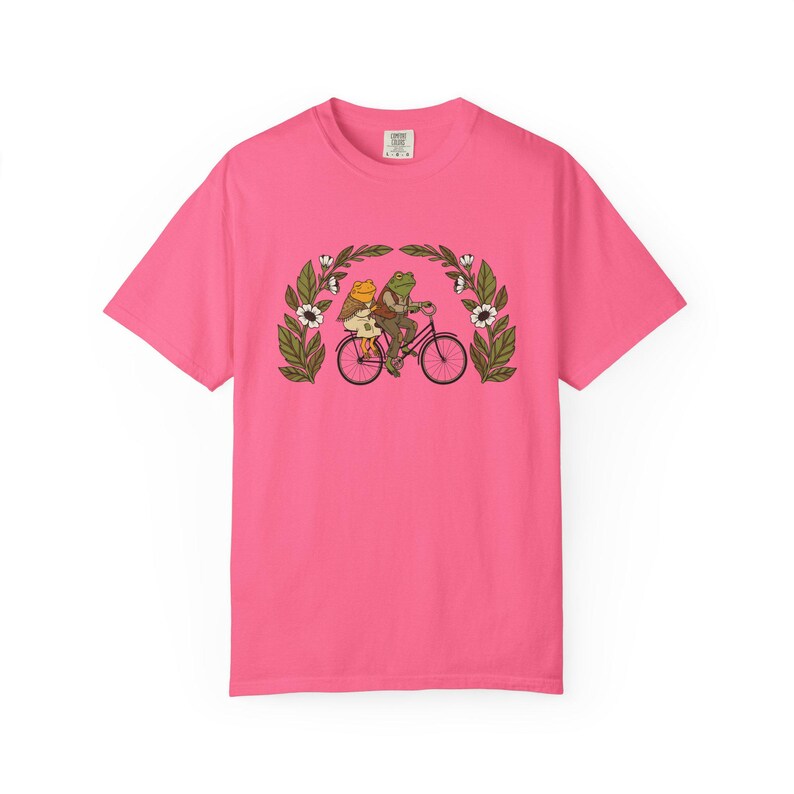 Puede incluir: Camiseta rosa brillante con una ilustraci&oacute;n caprichosa de dos ranas montando en bicicleta. Las ranas est&aacute;n rodeadas por un arco floral con flores blancas y hojas verdes. La camiseta es de color rosa s&oacute;lido.