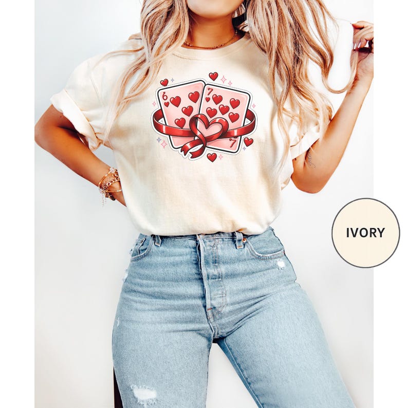 May include: Ivory-colored t-shirt featuring a graphic of two playing cards, a six and a seven of hearts, tied with a red ribbon. The shirt has a crew neck and short sleeves. The word "IVORY" is printed on a circle in the lower right corner.
