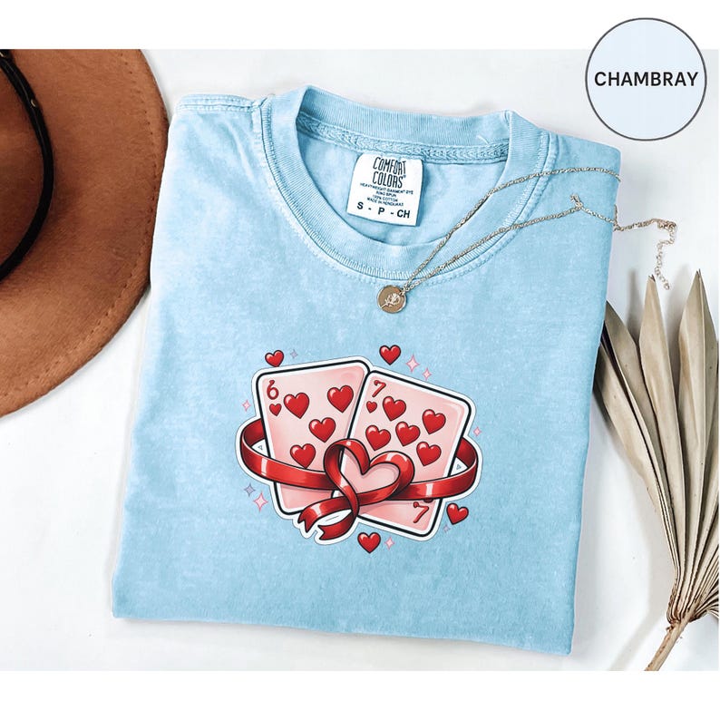 May include: A light blue t-shirt with a graphic of two playing cards, a six and a seven of hearts, wrapped in a red ribbon. The shirt has a Comfort Colors tag and a silver necklace. The word "CHAMBRAY" is in a circle in the upper right corner.