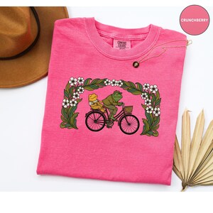 Puede incluir: Una camiseta rosa brillante Comfort Colors con un gr&aacute;fico l&uacute;dico de dos ranas montando en bicicleta. La imagen est&aacute; enmarcada dentro de un borde floral de flores blancas y follaje verde. La camiseta tiene cuello redondo y mangas cortas.