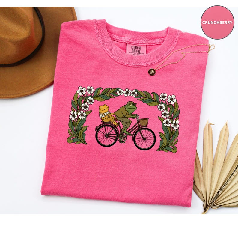 Puede incluir: Una camiseta rosa brillante Comfort Colors con un gr&aacute;fico l&uacute;dico de dos ranas montando en bicicleta. La imagen est&aacute; enmarcada dentro de un borde floral de flores blancas y follaje verde. La camiseta tiene cuello redondo y mangas cortas.
