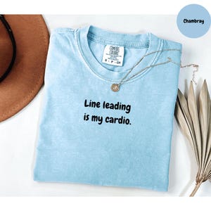 May include: Light blue t-shirt with the text "Line leading is my cardio." A silver necklace with a pendant is draped across the shirt. The shirt is labeled "Comfort Colors" and is size small.