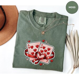 May include: A moss green t-shirt featuring a graphic of two playing cards with hearts, a red ribbon, and the numbers 6 and 7. A gold necklace and a brown hat are also visible.