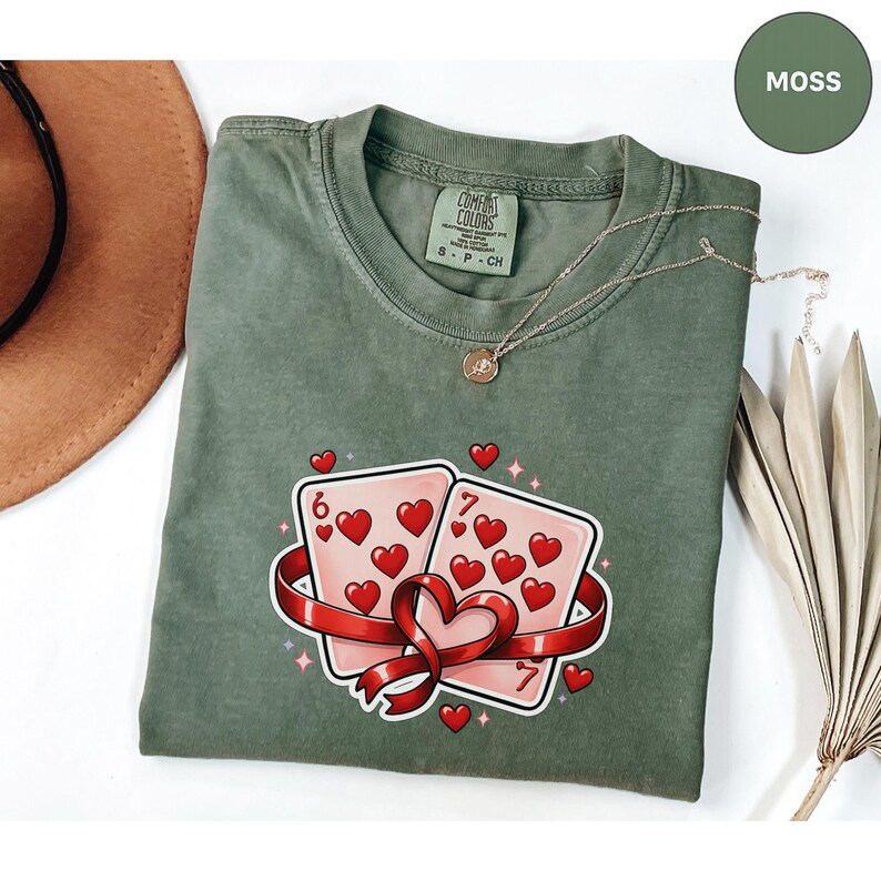 May include: A moss green t-shirt featuring a graphic of two playing cards with hearts, a red ribbon, and the numbers 6 and 7. A gold necklace and a brown hat are also visible.