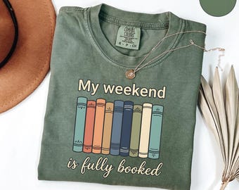 Op de afbeelding: Een mosgroen t-shirt met ronde hals en een afbeelding van negen kleurrijke boekruggen op een rij. Boven de boeken staat de tekst My weekend en onder de boeken staat de tekst is fully booked. Het t-shirt heeft een Comfort Colors label.