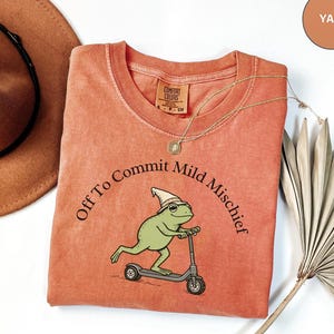 Könnte beinhalten: Ein yam-farbenes T-Shirt mit einer Grafik eines grünen Frosches, der einen spitzen Hut trägt und auf einem Roller fährt. Der Text "Off To Commit Mild Mischief" ist in einem schwarzen Bogen über dem Frosch gedruckt. Rundhalsausschnitt und Etikett.