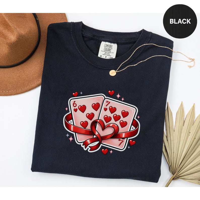 May include: Black t-shirt featuring a graphic of two playing cards with hearts, a red ribbon, and the numbers 6 and 7. The shirt has a crew neck and a tag that says "Comfort Colors". A gold necklace and a brown hat are also visible.
