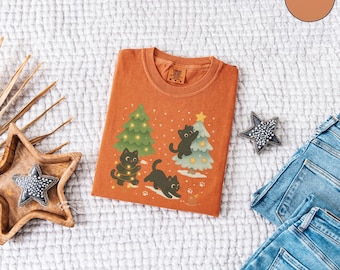 Puede incluir: Una camiseta naranja con un diseño navideño con gatos negros y árboles de Navidad decorados. La camiseta está colocada sobre una superficie blanca texturizada, junto a una bandeja de madera en forma de estrella con una decoración en forma de estrella y unos vaqueros.