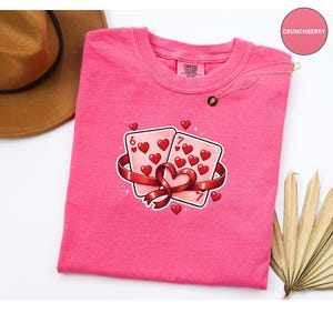 May include: A pink Comfort Colors t-shirt featuring a graphic of two playing cards with hearts, a red ribbon, and small hearts. The cards are a six and a seven. A gold necklace and a brown hat are also visible.