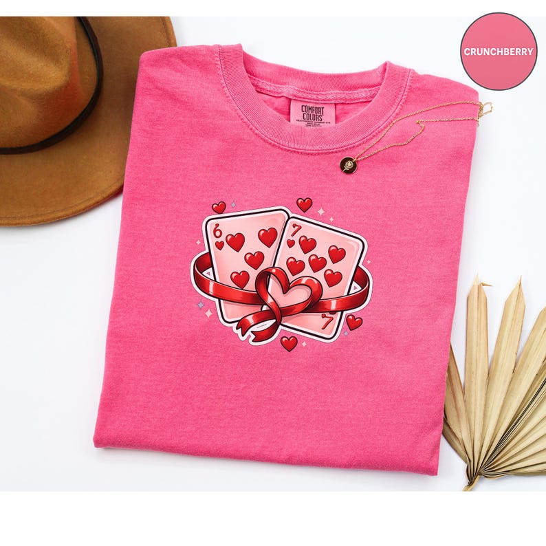 May include: A pink Comfort Colors t-shirt featuring a graphic of two playing cards with hearts, a red ribbon, and small hearts. The cards are a six and a seven. A gold necklace and a brown hat are also visible.