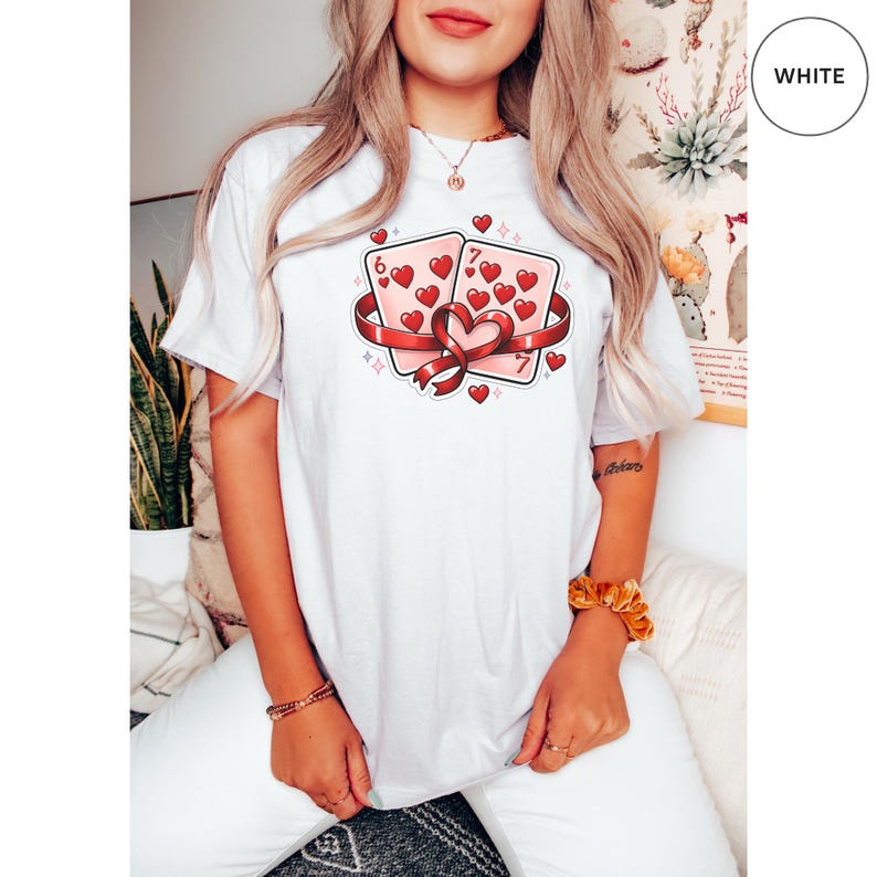 May include: White t-shirt featuring a graphic of two playing cards, a six and a seven of hearts, wrapped with a red ribbon. The cards are filled with red hearts. The word "WHITE" is in a white circle in the upper right corner.
