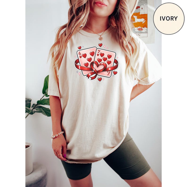 May include: Ivory-colored t-shirt featuring a graphic of two playing cards with hearts, a red ribbon, and the numbers 6 and 7. The design includes small hearts and sparkles. The shirt is a relaxed fit.