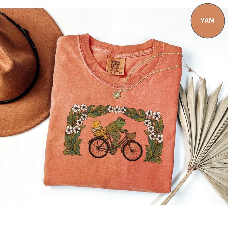 Puede incluir: Una camiseta de color coral con una ilustraci&oacute;n caprichosa de dos ranas montando en bicicleta a trav&eacute;s de un arco floral. La camiseta tiene cuello redondo y mangas cortas. El texto "COMFORT COLORS" es visible en la etiqueta.