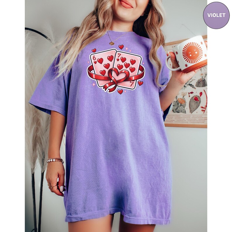 May include: A violet-colored t-shirt featuring a graphic of two playing cards with hearts and a ribbon. The cards are pink with red hearts and the numbers 6 and 7. The shirt is a comfortable, oversized fit.