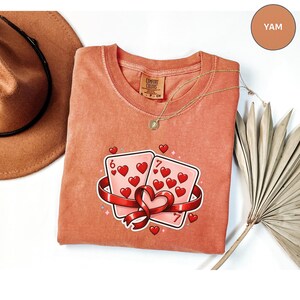 May include: A coral-colored t-shirt with a graphic of two playing cards, a six and a seven of hearts, wrapped in a red ribbon. A gold necklace and a brown hat are also visible.